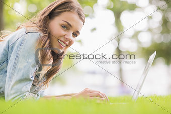 Happy student lying on the grass using her laptop looking at camera