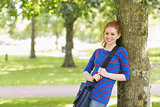 Smiling redhead student leaning against a tree