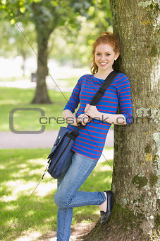 Happy redhead student leaning against a tree