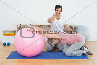 Therapist gesturing thumbs up by senior woman with yoga ball