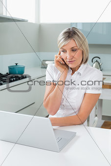 Casual woman using laptop while on call in kitchen