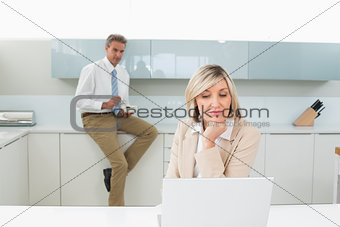 Woman using laptop and man in background at kitchen