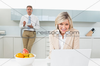 Woman using laptop and man in background at kitchen
