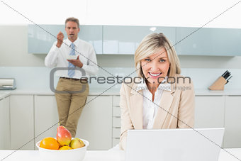 Smiling woman with laptop and man in background at kitchen