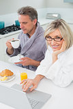 Couple in bathrobes with coffee and juice using laptop in kitchen
