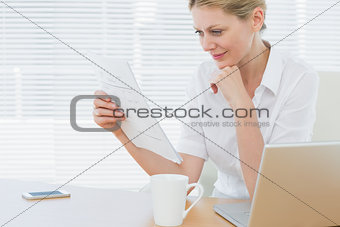 Businesswoman reading a document besides laptop at desk