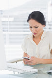 Businesswoman text messaging in front of computer at desk
