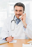 Smiling male doctor using phone while writing notes