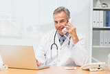 Smiling doctor using laptop and talking on phone at desk
