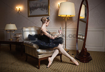 Ballerina in black tutu sitting in armchair in front of mirror and taking self portrait with camera on her cell phone