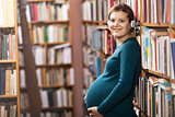Young pregnant woman in headphones looking at camera while standing in library