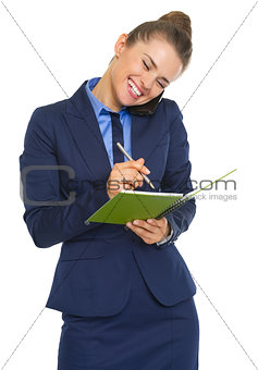 Happy business woman talking mobile phone and writing in notepad