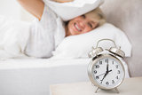 Woman covering ears with pillow with alarm clock in foreground