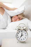Woman covering ears with pillow with alarm clock in foreground