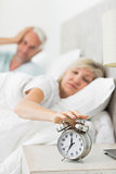 Mature woman extending hand to alarm clock in bed