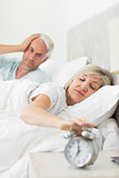 Man covering ears while woman extending hand to alarm clock in bed