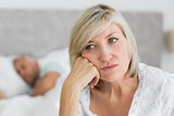 Tensed mature woman sitting in bed with man in background