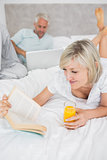 Couple reading book and using laptop in bed