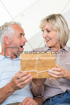 Smiling woman surprising mature man with a gift on sofa