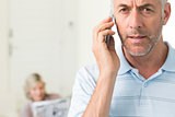 Mature man using cellphone with woman reading newspaper in background