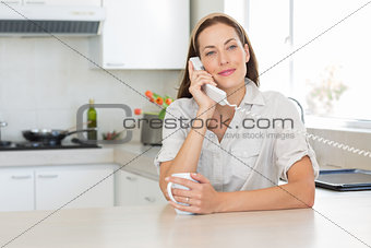 Smiling woman with coffee cup using landline phone in kitchen