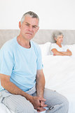 Angry senior man on bed with woman in background