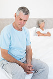 Sad senior man on bed with wife in background