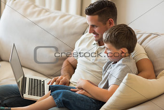 Relaxed father and son using laptop on sofa