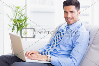 Businessman sitting on couch using his laptop smiling at camera