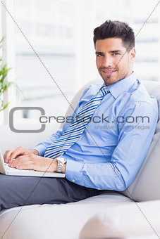 Businessman on couch using his laptop smiling at camera