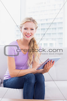 Happy blonde sitting on her sofa using her tablet pc
