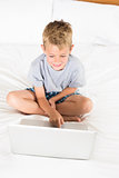 Cheerful blonde boy sitting on bed using laptop