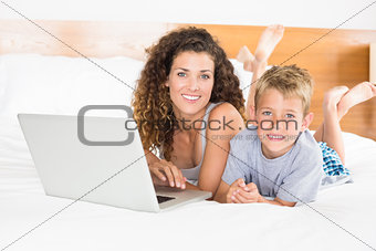 Smiling blonde boy and mother lying on bed using laptop