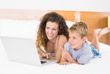 Cheerful blonde boy and mother lying on bed using laptop