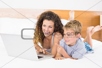 Cheerful blonde boy and mother lying on bed using laptop