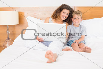 Happy mother and son sitting on bed using laptop