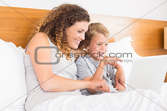 Smiling mother and son sitting on bed looking at laptop