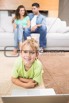 Happy little boy using laptop on the rug with parents sitting sofa