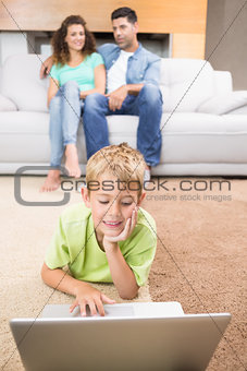 Cute little boy using laptop on the rug with parents sitting sofa