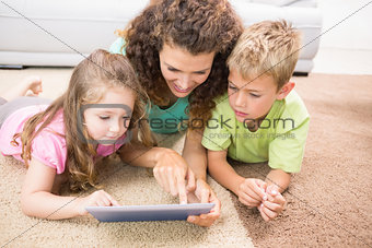 Happy siblings lying on the rug using tablet with their mother