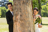 Happy newlywed couple behind tree trunk in park
