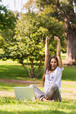 Cheerful woman raising hands with laptop in park