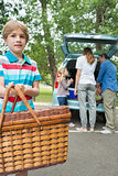Boy with picnic basket while family in background at car trunk