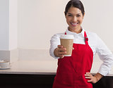 Pretty young barista offering cup of coffee to go smiling at camera