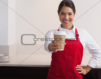 Pretty young barista offering cup of coffee to go smiling at camera