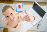 Happy young woman with books and laptop in kitchen showing thumb