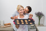 Man kissing woman's cheek as she holds freshly baked cookies in kitchen