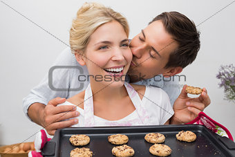 Man kissing woman's cheek as she holds freshly baked cookies in kitchen