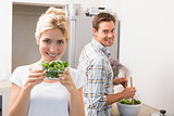 Woman holding bowl of leaves with man preparing salad in background in kitchen