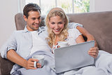 Relaxed couple with coffee cups using laptop on sofa